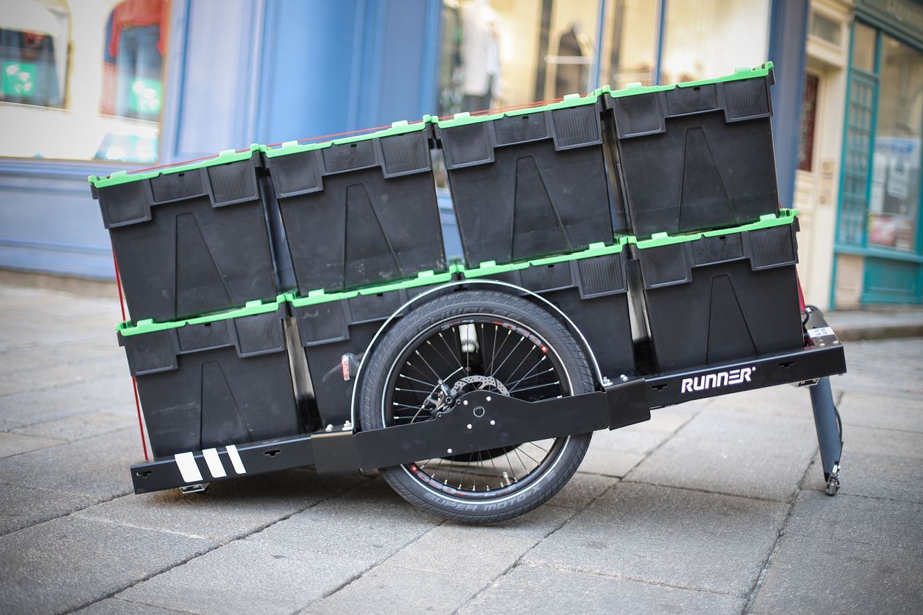 Ein FlexiModal Anhänger mit schwarzen Kisten beladen steht abgekoppelt vom Fahrrad auf einem Platz.